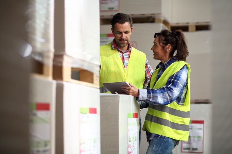 Two workers are wearing neon yellow safety vests. One worker is holding a clipboard. There are boxes in the foreground. The workers are in a warehouse